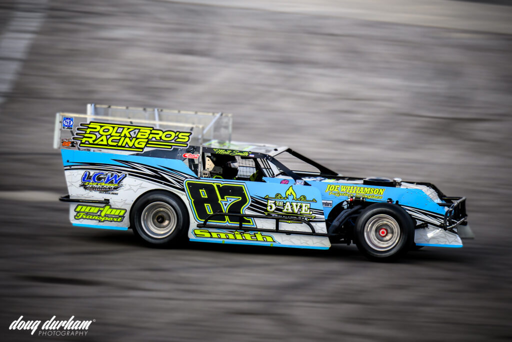 A blue and black Polk Bros race car with the number 87 on the side is speeding at the Speedrome. The vehicle features various sponsor logos and is captured in motion.