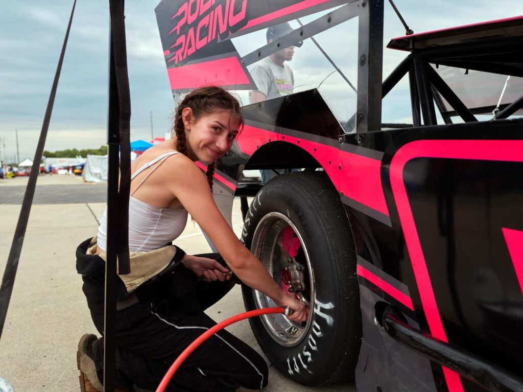 Racer Gillian Leppado is crouched next to a racing car with bold pink and black graphics. She is inflating the tire using an air hose. The sky is overcast, and other cars and people are visible in the background.