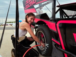 Racer Gillian Leppado is crouched next to a racing car with bold pink and black graphics. She is inflating the tire using an air hose. The sky is overcast, and other cars and people are visible in the background.