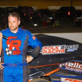 A man in a blue racing suit with red and black design stands beside a race car labeled "Polk Bro's Racing" and "Mellor's Performance." He is being interviewed by another man holding a microphone. People and cars are visible in the background, with a woman sitting on a wall.