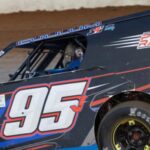 A modified race car with number 95 on the side races on a dirt track. It features sponsor logos and blue, black, red, and white markings. The driver's helmet is partially visible through the window, and the car leans slightly on the track, highlighting its speed and motion.