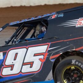 A modified race car with number 95 on the side races on a dirt track. It features sponsor logos and blue, black, red, and white markings. The driver's helmet is partially visible through the window, and the car leans slightly on the track, highlighting its speed and motion.