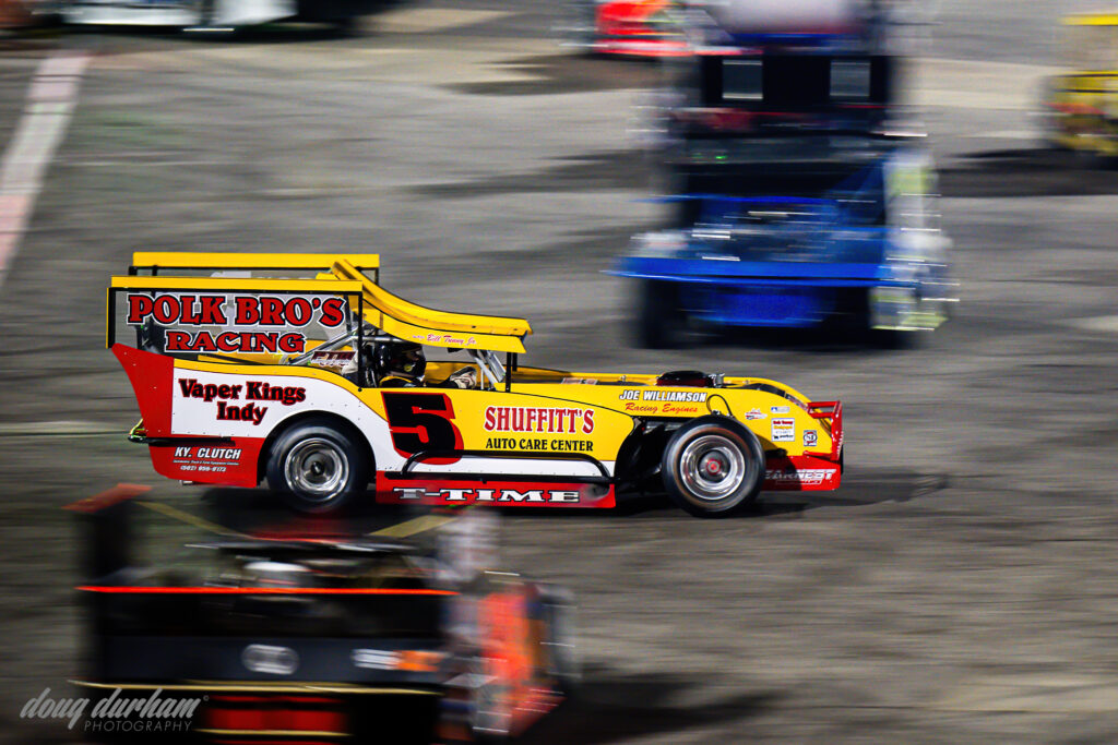 A vintage-style race car with "Polk Bro's Racing" is speeding on a racetrack. The car is predominantly red and yellow, with the number 5 prominently displayed. Other blurred vehicles are visible, implying speed.