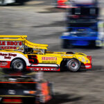 A vintage-style race car with "Polk Bro's Racing" is speeding on a racetrack. The car is predominantly red and yellow, with the number 5 prominently displayed. Other blurred vehicles are visible, implying speed.