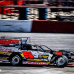 A race car with "Polk Bro's Racing" is speeding on a racetrack. The car is predominantly red and yellow, with the number 5 prominently displayed. Other blurred vehicles are visible, implying speed.