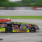 A race car with "Polk Bro's Racing" and "Shuffit's Auto Care Center" on the side is speeding on a racetrack. The car is predominantly red and yellow, with the number 5 prominently displayed. Other blurred vehicles are visible, implying speed.