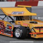 A bright yellow and orange modified racing car is on a track, featuring a large rear wing and various sponsor logos. The car is number 55. The track has red and white barriers in the background.