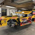 A yellow and black race car is parked in a garage. The car has the number 5 prominently displayed. The garage is filled with tools and equipment, with a wooden ceiling and white walls.