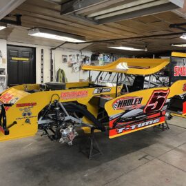 A yellow and black race car is parked in a garage. The car has the number 5 prominently displayed. The garage is filled with tools and equipment, with a wooden ceiling and white walls.