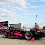 Race car with "Pork Bro's Racing" in pink lettering on the side, featuring a bold pink and black design. The car has large rear spoilers and specialized tires. People are in the background, near a trailer and equipment, under a blue sky with scattered clouds.
