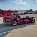 Race car with "Pork Bro's Racing" in pink lettering on the side, featuring a bold pink and black design. The car has large rear spoilers and specialized tires. People are in the background, near a trailer and equipment, under a blue sky with scattered clouds.