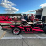 Race car with "Pork Bro's Racing" in pink lettering on the side, featuring a bold pink and black design. The car has large rear spoilers and specialized tires. People are in the background, near a trailer and equipment, under a blue sky with scattered clouds.