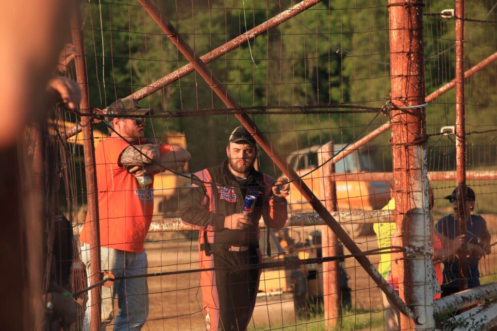 Racer John Walp in a racing suit stands behind a wire fence, holding a drink can, smiling, and leaning on the fence. Another man in an orange shirt stands beside him, wearing a cap and sunglasses. Construction vehicles and blurred greenery are in the background. The scene is lit by warm sunlight.