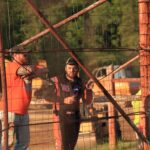 Racer John Walp in a racing suit stands behind a wire fence, holding a drink can, smiling, and leaning on the fence. Another man in an orange shirt stands beside him, wearing a cap and sunglasses. Construction vehicles and blurred greenery are in the background. The scene is lit by warm sunlight.