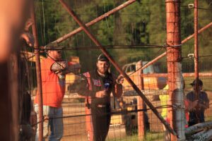 Racer John Walp in a racing suit stands behind a wire fence, holding a drink can, smiling, and leaning on the fence. Another man in an orange shirt stands beside him, wearing a cap and sunglasses. Construction vehicles and blurred greenery are in the background. The scene is lit by warm sunlight.