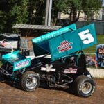 A teal sprint car, marked with the number 5, races on a dirt track. The car features prominent logos, and its tires kick up dust. In the background, spectators sit on blue bleachers, and a few seated on a bench watch the race. Trees and a fence line the track.