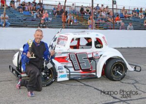 Tim Skinner kneels on the racetrack, holding a trophy and a checkered flag. Beside him is his white race car with the number 15 and various sponsor logos. People are seated in the bleachers in the background.