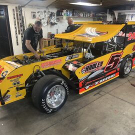 A yellow and black race car is parked in a garage. The car has the number 5 prominently displayed. The garage is filled with tools and equipment, with a wooden ceiling and white walls.