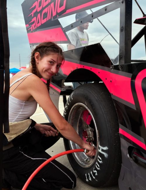 Racer Gillian Leppado is crouched next to a racing car with bold pink and black graphics. She is inflating the tire using an air hose. The sky is overcast, and other cars and people are visible in the background.