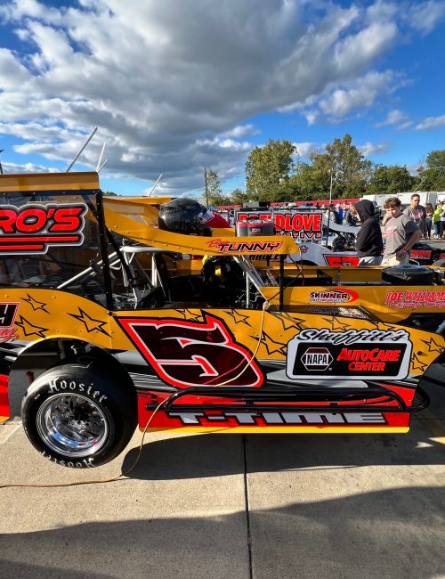 Features racer Ben Tunny's bright yellow and red racing car with the number 5 on its side, featuring various sponsor logos. The car has large chrome wheels. People are in the background on a sunny day with a few clouds in the sky.