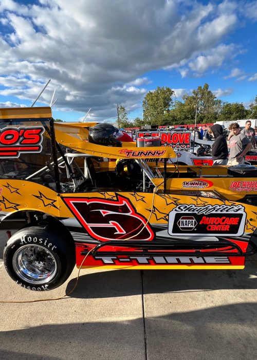 Features racer Ben Tunny's bright yellow and red racing car with the number 5 on its side, featuring various sponsor logos. The car has large chrome wheels. People are in the background on a sunny day with a few clouds in the sky.