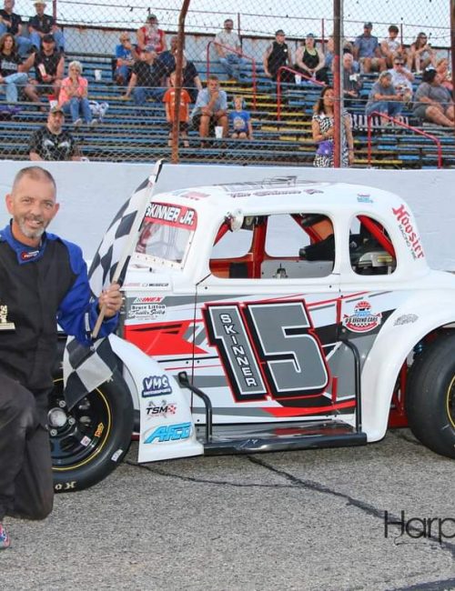 Tim Skinner kneels on the racetrack, holding a trophy and a checkered flag. Beside him is his white race car with the number 15 and various sponsor logos. People are seated in the bleachers in the background.