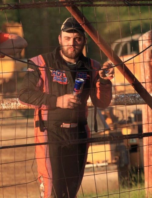 Racer John Walp in a racing suit stands behind a wire fence, holding a drink can, smiling, and leaning on the fence. Another man in an orange shirt stands beside him, wearing a cap and sunglasses. Construction vehicles and blurred greenery are in the background. The scene is lit by warm sunlight.