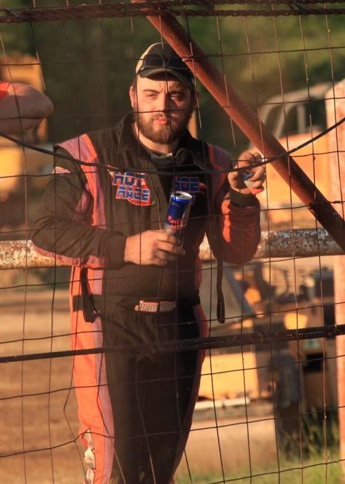 Racer John Walp in a racing suit stands behind a wire fence, holding a drink can, smiling, and leaning on the fence. Another man in an orange shirt stands beside him, wearing a cap and sunglasses. Construction vehicles and blurred greenery are in the background. The scene is lit by warm sunlight.
